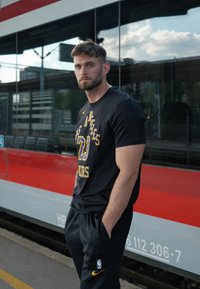 Svart T-skjorte med korte ermer og gul "Los Angeles Lakers 23" trykk, kombinert med svarte bukser. Bakgrunnen har et rødt og grått tog.