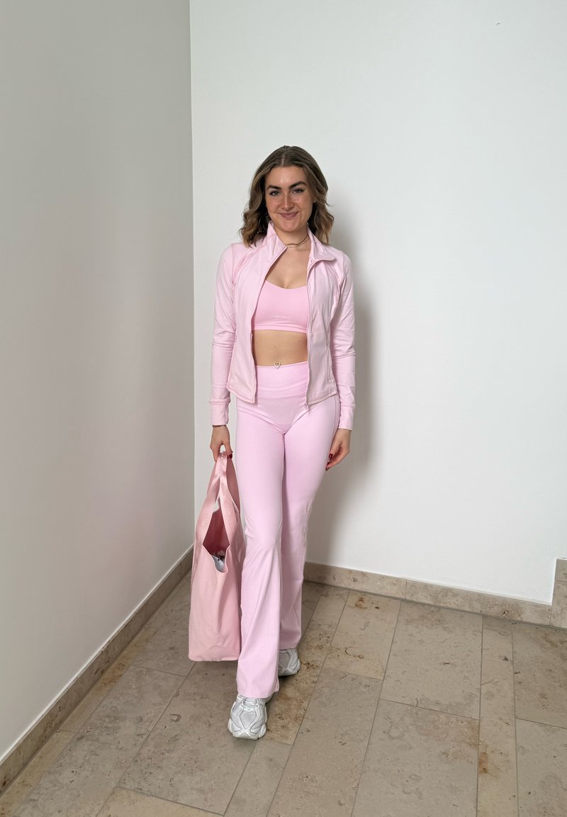 Donna che indossa un completo sportivo rosa chiaro tiene una borsa rosa abbinata, in piedi su un pavimento piastrellato davanti a una parete bianca.