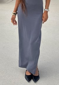 Maxikleid in Grau, enganliegend aus glattem Stoff, kombiniert mit schwarzen spitzen Schuhen. Accessoires umfassen mehrere Armbänder und eine Uhr.