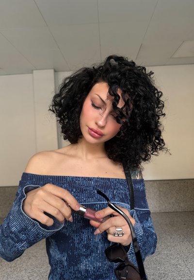 Top azul de hombros al descubierto con patrón texturizado; sosteniendo un brillo labial transparente con tapa plateada; gafas de sol negras en la otra mano; cabello rizado negro.