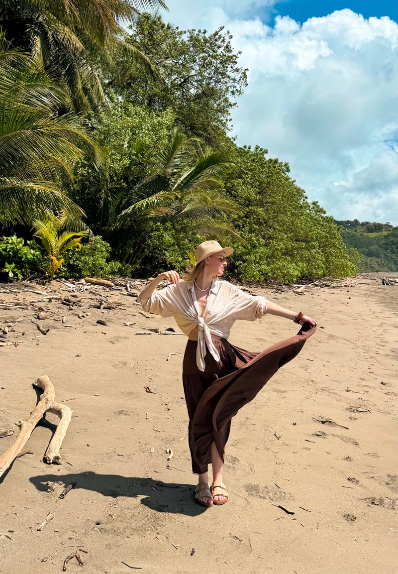 Blouse beige clair nouée, jupe longue marron, chapeau à large bord et sandales. En arrière-plan, une plage de sable et une végétation verte luxuriante.