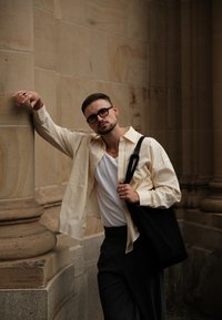 Hellbeige, gestreifte Hemd über einem weißen T-Shirt, schwarze Hose und eine schwarze Tragetasche. Das Model steht neben einer Steinsäule und zeigt eine entspannte Pose.