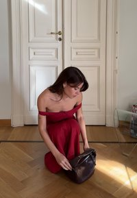 Czarna skórzana torebka z zapięciem na zakładkę i zamkiem błyskawicznym, trzymana przez osobę w czerwonej sukience z odkrytymi ramionami, siedzącą na drewnianej podłodze.