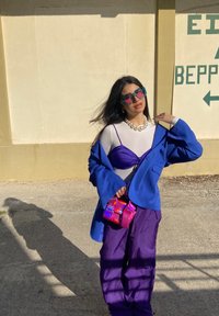 Mulher vestindo um blazer azul oversized, calças roxas, camiseta branca, colar de pérolas chunky, óculos de sol azul-turquesa, segurando uma bolsa iridescente rosa do lado de fora ao sol.