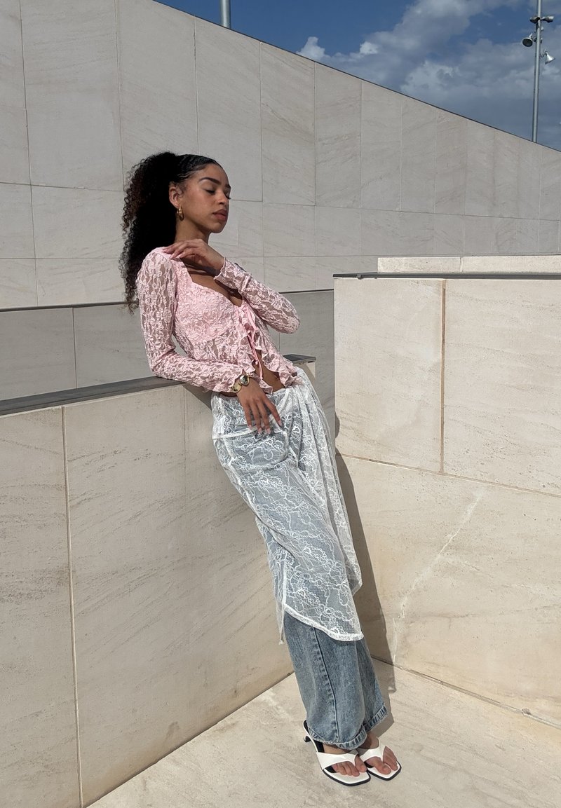 Femme aux cheveux bouclés appuyée contre un mur de pierre claire, portant un haut en dentelle rose, une superposition translucide en dentelle blanche, un jean bleu et des sandales blanches à bout carré.