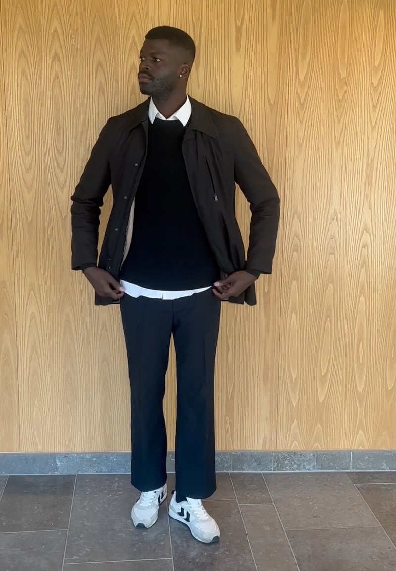 Homme portant une veste noire, un pull noir sur une chemise blanche, un pantalon noir et des baskets blanches, debout contre un mur en bois lambrissé en ajustant sa veste.