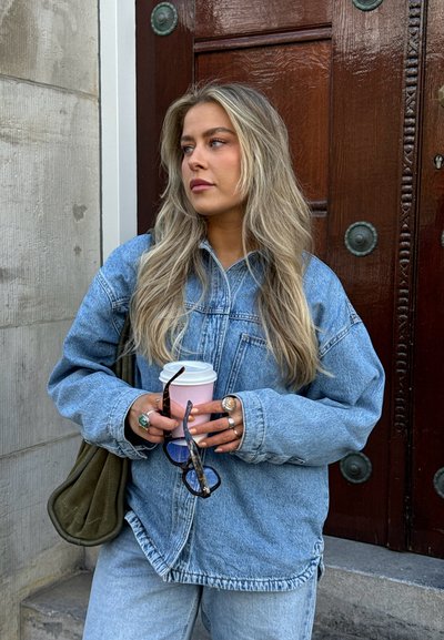 Chaqueta de denim y jeans en azul claro, con un corte relajado. Sosteniendo una taza rosa y gafas de sol con montura negra, adornada con anillos.