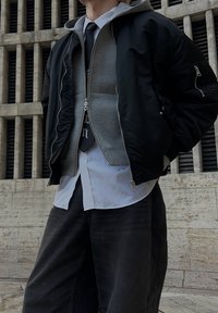 Personne portant une veste bomber noire superposée, un sweat à capuche gris, une chemise blanche, une cravate noire et un jean foncé, debout devant un bâtiment en pierre avec des fenêtres verticales.