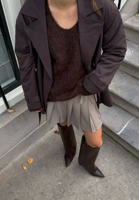 Hnědý dlouhý kabát, huňatý tmavý svetr, plisovaná béžová sukně a hnědé kozačky s špičatou špičkou sahající ke kolenům. Jsou viditelné detaily texturované látky.