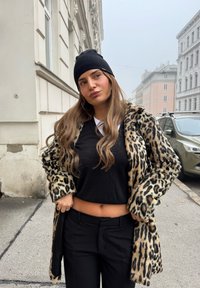 Fuskpälsjacka med leopardmönster, svart tröja och svarta byxor med hög midja. Modellen poserar utomhus, med långt och vågigt hår, bär en svart mössa.