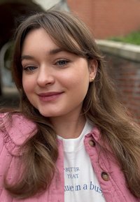 Jonge vrouw met lang bruin haar, die kleine gouden oorbellen draagt, een roze gestructureerde jas en een wit shirt met blauwe tekst, buiten bij een bakstenen muur.
