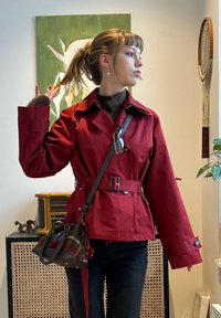 Femme avec queue de cheval portant une veste rouge ceinturée et un pantalon sombre, tenant ses cheveux, avec une œuvre d'art et une radio vintage en arrière-plan.