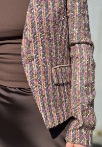 Giacca in tweed multicolore con bottone dorato sopra un top marrone a coste e una gonna marrone liscia, mostrando la dettagliata trama del tessuto e delle maniche.