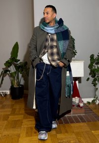 Manteau gris, chemise à carreaux, t-shirt blanc, jean foncé, écharpe à carreaux bleu-vert, détail chaîne argentée, baskets avec accents réfléchissants.