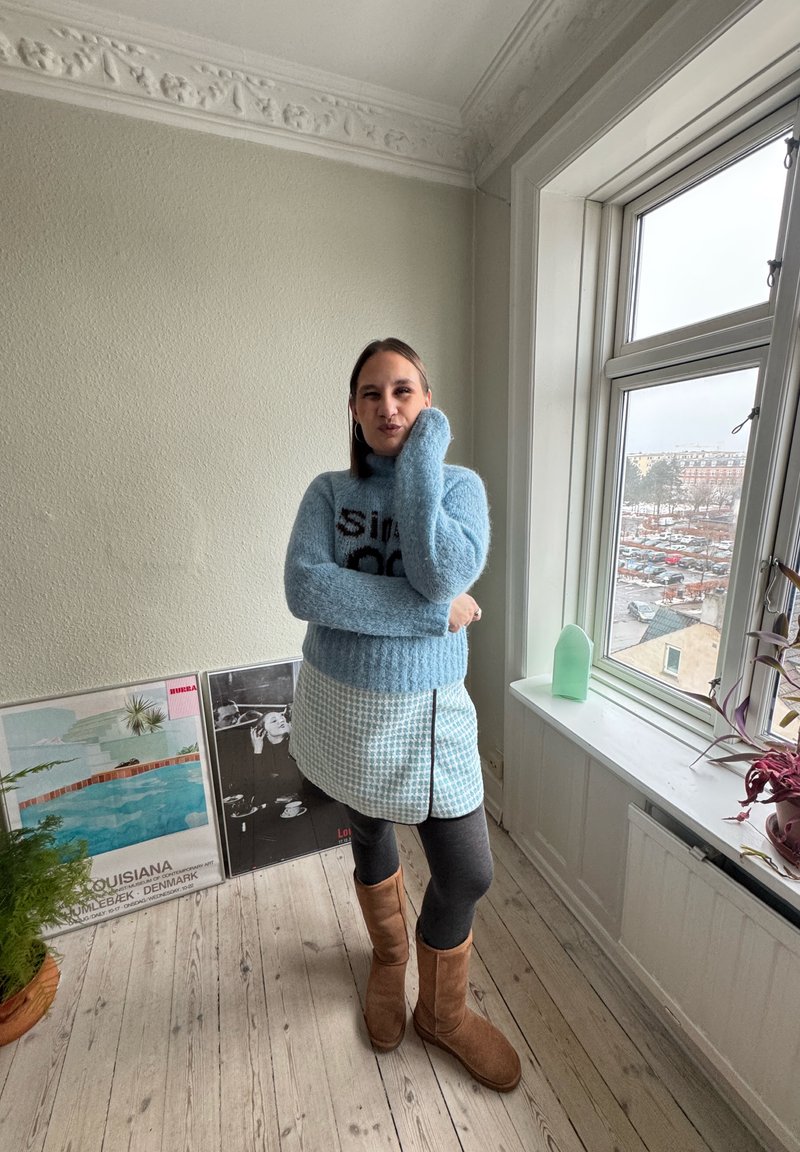 Femme en pull bleu, jupe à motifs et bottes marron, debout près d'une fenêtre avec des plantes et des affiches encadrées, sur un sol en bois.