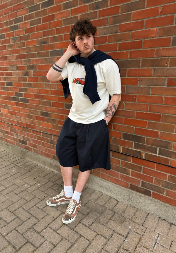 Jeune homme aux cheveux bouclés et tatouages, portant un t-shirt blanc avec imprimé voiture, un short noir, des baskets marron et un pull bleu marine posé sur les épaules.