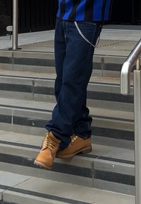 Rjave usnjene delovne čevlje z rumenimi vezalkami, oblečene s temno modrimi jeans hlačami, postavljene na sivo in črno črtaste stopnice. Vidna je verižna dekoracija.