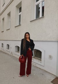 Schwarzer übergroßer Blazer, schwarzes Oberteil, weite bordeauxrote Hose, rote Handtasche und Riemchensandalen. Lange geflochtene Haare und sichtbare Ohrringe.
