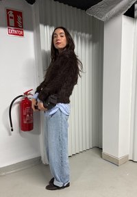 Femme en veste en fourrure marron et jeans debout à côté d'un extincteur rouge fixé sur un mur blanc dans un intérieur industriel.
