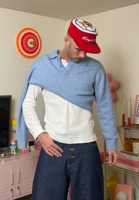Hellblauer, gestrickter Pullover über einem weißen Langarmshirt, kombiniert mit dunklerjeans. Rote Baseballkappe mit Logo.