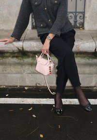 Persona seduta su una panchina di pietra, indossando un cardigan grigio a maglia, pantaloni neri, collant neri a pois, mentre tiene in mano una piccola borsa rosa con un ciondolo di perle colorate.