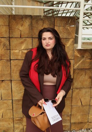 Saco marrón sobre una blusa beis, bufanda roja y falda marrón. Ella sostiene unas gafas de sol y un bolso beige con una etiqueta. Fondo de madera texturizada.