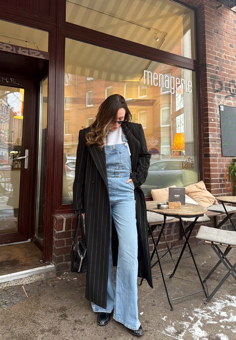 Donna con cappotto nero a righe sopra una salopette di jeans, in piedi fuori da un caffè con posti a sedere all'aperto e pareti di mattoni.