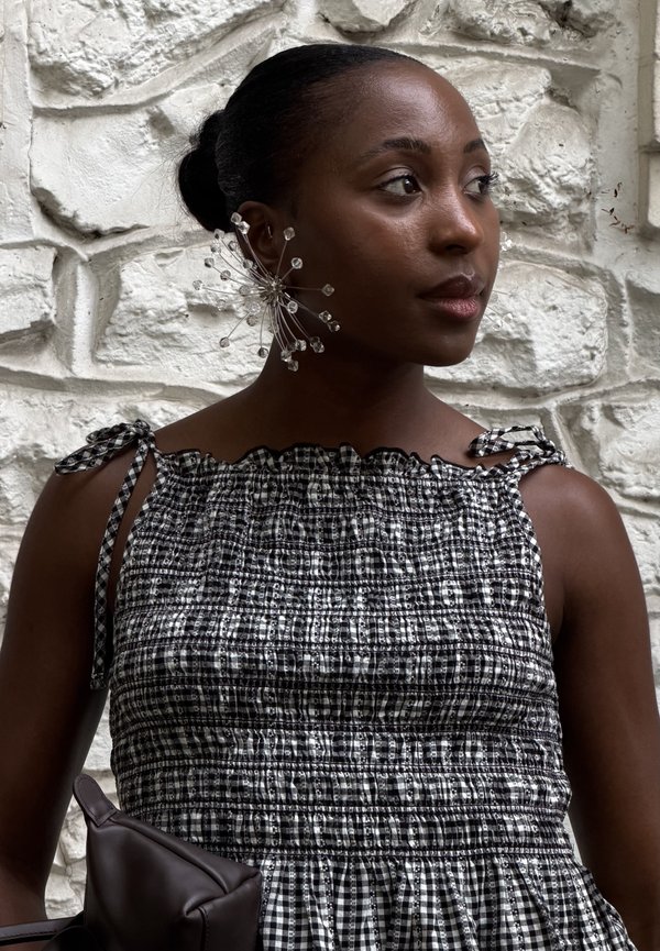 Femme à la peau foncée portant une robe à carreaux noire et blanche et une grande boucle d'oreille décorative, debout contre un mur de pierre blanche texturé.