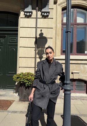 Abrigo gris de doble botonadura en mezcla de lana, cerrado con un cinturón negro, usado sobre pantalones negros; de pie junto a una puerta verde y una planta en maceta.