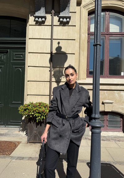 Abrigo gris de doble botonadura en mezcla de lana, cerrado con un cinturón negro, usado sobre pantalones negros; de pie junto a una puerta verde y una planta en maceta.