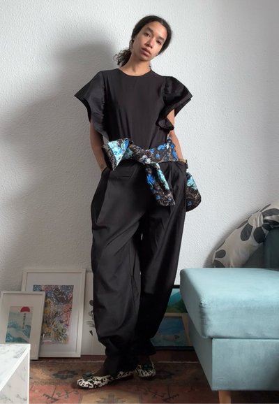 Mujer de pie en el interior vistiendo una blusa negra con mangas con volantes, pantalones negros holgados, chaqueta floral anudada a la cintura y zapatos estampados.