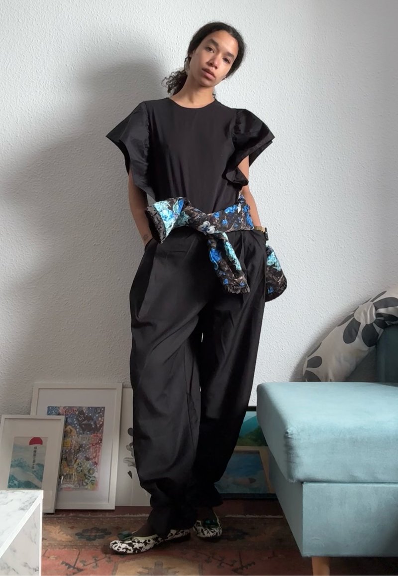 Femme debout à l'intérieur portant un haut noir à manches volantées, un pantalon noir ample, une veste fleurie nouée à la taille et des chaussures à motifs.