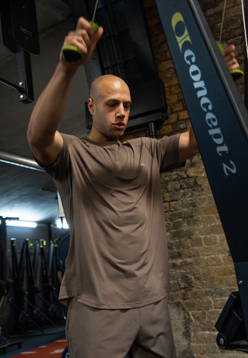 Homme portant des vêtements de sport marron utilisant un ergomètre Concept 2 dans une salle de sport avec des murs en briques.