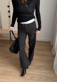 Personne portant un haut noir à manches longues, un pantalon noir taille haute avec ceinture, des bottes noires, et portant un sac à main noir sur un sol en bois à l'intérieur.
