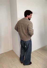 Homme aux cheveux bouclés et barbe, portant un pull rayé beige et un jean bleu ample, debout face à un mur blanc sur un sol en bois clair.