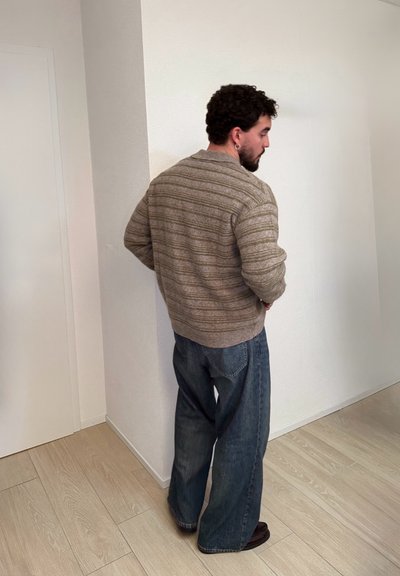 Hombre con cabello rizado y barba, vestido con un suéter beige con rayas y jeans azules holgados, de pie frente a una pared blanca sobre un suelo de madera clara.