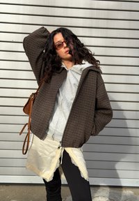 Femme aux longs cheveux bouclés portant une veste marron à carreaux, un sweat à capuche gris, une jupe crème ornée de dentelle, des leggings noirs, des lunettes de soleil marron et un sac à bandoulière.