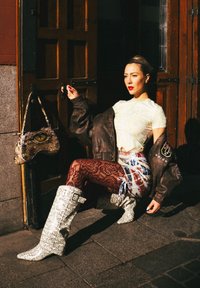 Braune übergroße Lederjacke, weißes gemustertes Oberteil, rote gemusterte Leggings und weiße kniehohe Stiefel. Handtasche mit Augenmuster. Sonnendurchflutete Straßenszene.