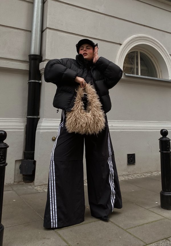 Zwarte pufferjack, oversized zwarte broek met witte zijkanten, en een pluizige beige tas. Buitensetting met een grijze muur en stoep.