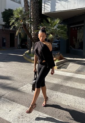 Mujer con top negro de un solo hombro y leggings capri caminando por un paso de peatones urbano soleado, llevando un bolso negro, con palmeras de fondo.