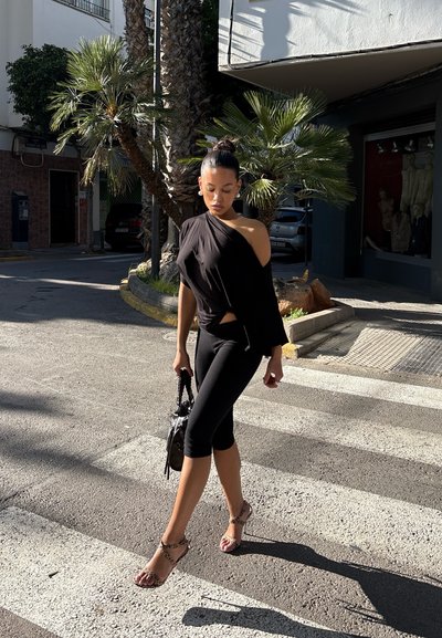 Mujer con top negro de un solo hombro y leggings capri caminando por un paso de peatones urbano soleado, llevando un bolso negro, con palmeras de fondo.