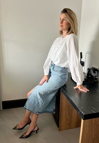 Fehér, texturált blúz puffos ujjakkal, világoskék farmer közép hosszú szoknyával és fekete, hegyes orrú magassarkúval, arany kiegészítőkkel.