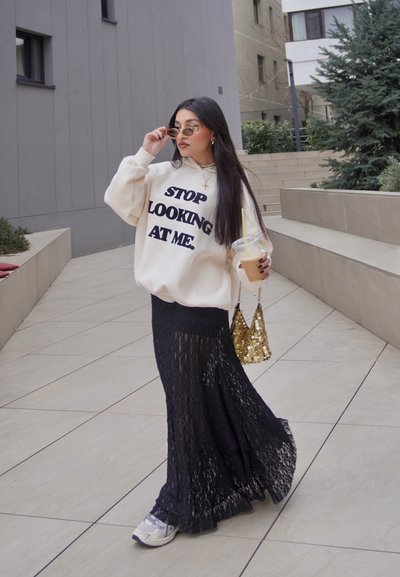 Sudadera crema con texto negro en negrita, falda larga de encaje negra, collar con cruz de plata, gafas de sol y un bolso de lentejuelas doradas.