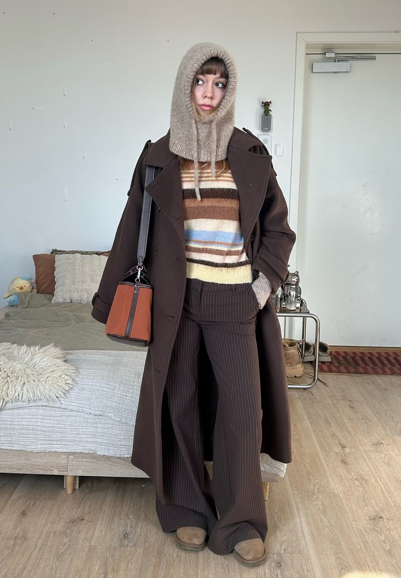 Frau mit braunem Mantel, gestreiftem Pullover, Nadelstreifenhose, gestrickter Kapuze und brauner Handtasche, stehend in einem Schlafzimmer.