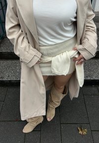 Beige trenchcoat over en hvid top, parret med en cremet struktureret miniskirt bundet i taljen og knæhøje lysebrune ruskind støvler.