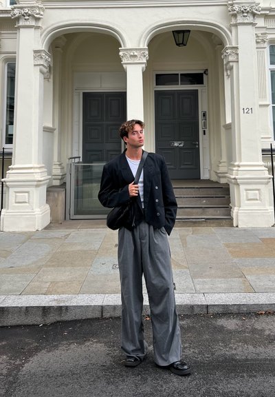 Blazer negro de doble botonadura, camiseta blanca, pantalones anchos grises y zapatos negros. El fondo presenta una arquitectura clásica con columnas.