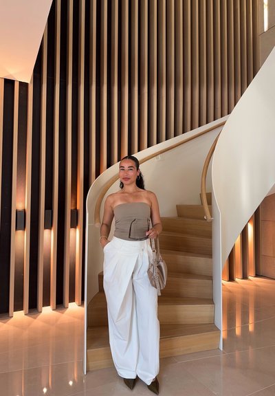 Top strapless de color beige claro combinado con pantalones blancos de pierna ancha. Tacones puntiagudos en verde oliva oscuro. Escalera de madera en espiral y pared de madera texturizada en el fondo.