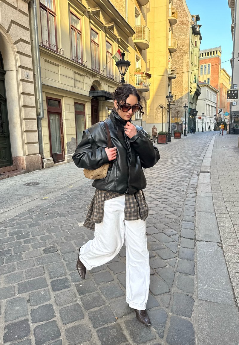 Donna con giacca di pelle nera, pantaloni bianchi e occhiali da sole che cammina su una strada acciottolata fiancheggiata da edifici storici sotto una luce solare soffusa.