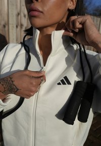 Personne portant une veste blanche Adidas en train de dézippper le col, tenant des poignées de corde à sauter noires, en extérieur avec une clôture en bois en arrière-plan.