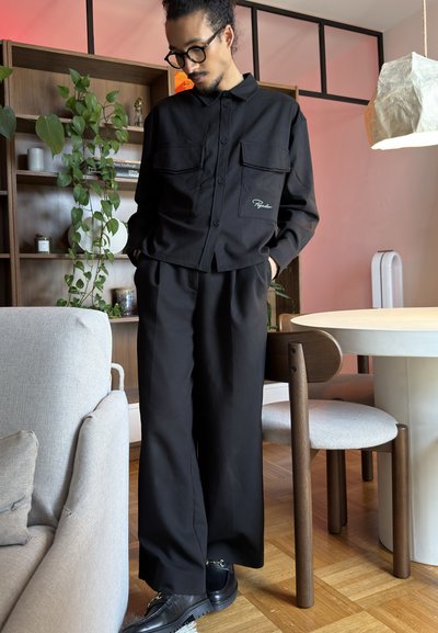 Hombre con camisa negra abotonada y pantalones de pierna ancha de pie en una habitación moderna con estanterías de madera, plantas y mesa de comedor.